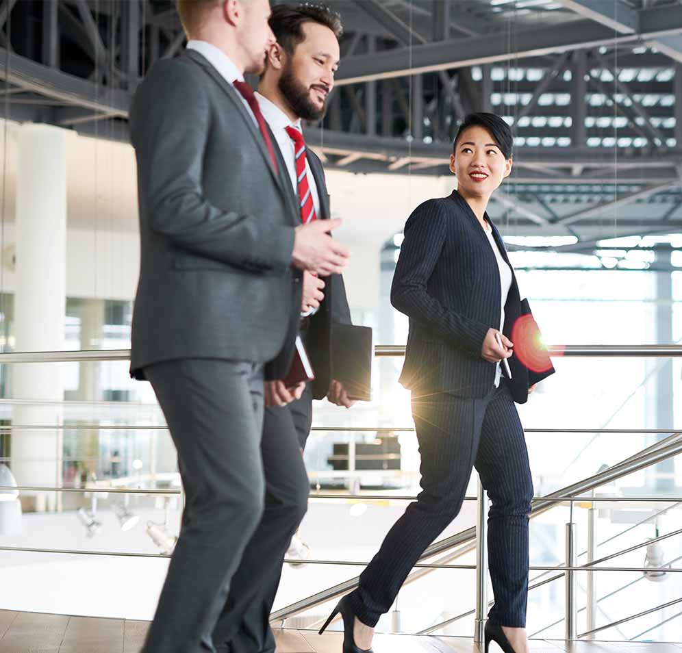 Three professionals walking in a modern office building, engaged in a conversation. The woman in the foreground looks back with a smile at the two men following her, who are dressed in suits and carrying documents. The setting suggests a dynamic and collaborative work environment.